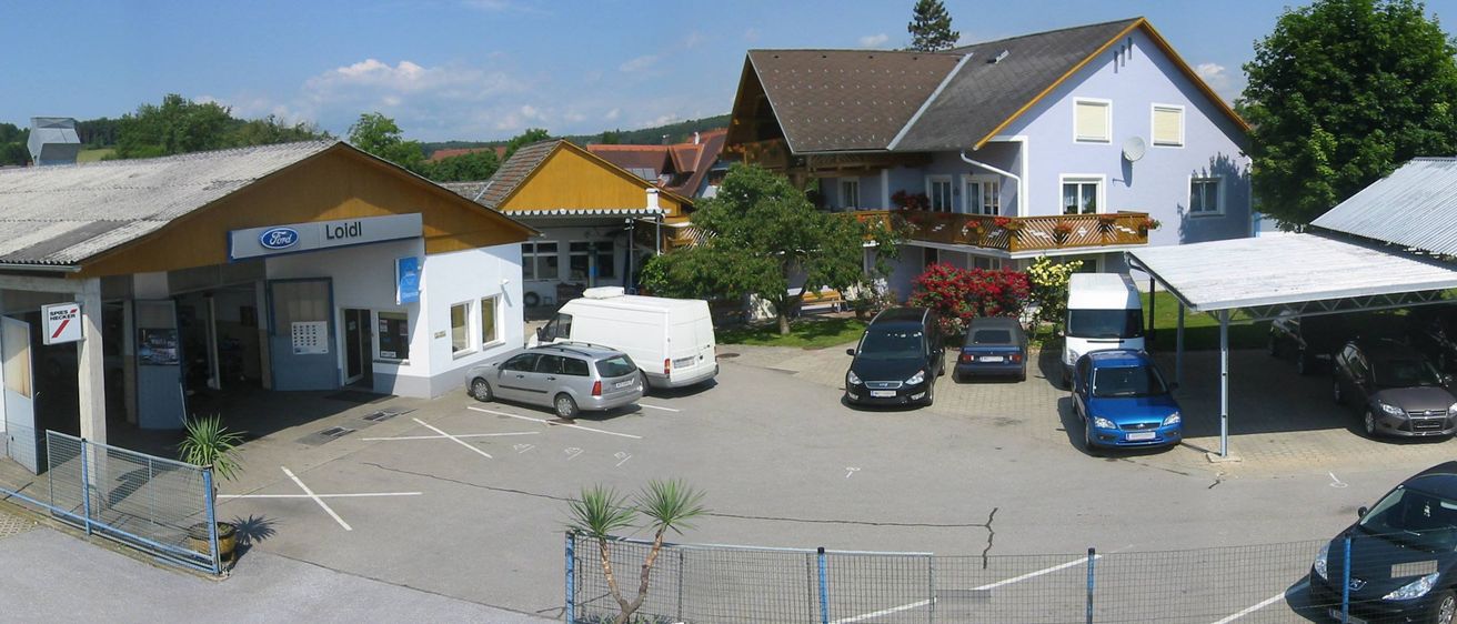 Ein Blick von oben auf einen Parkplatz mit mehreren Autos, die vor einem Haus mit Balkon und roten Blumen an den Geländern geparkt sind.