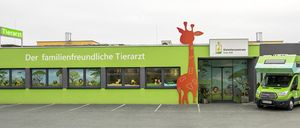 Eine Tierarztpraxis mit grünen Wänden und einer Giraffendekoration vor dem Gebäude. Das Schild steht 'freundlicher Tierarzt'. Drei Fenster zeigen Tierbilder.
