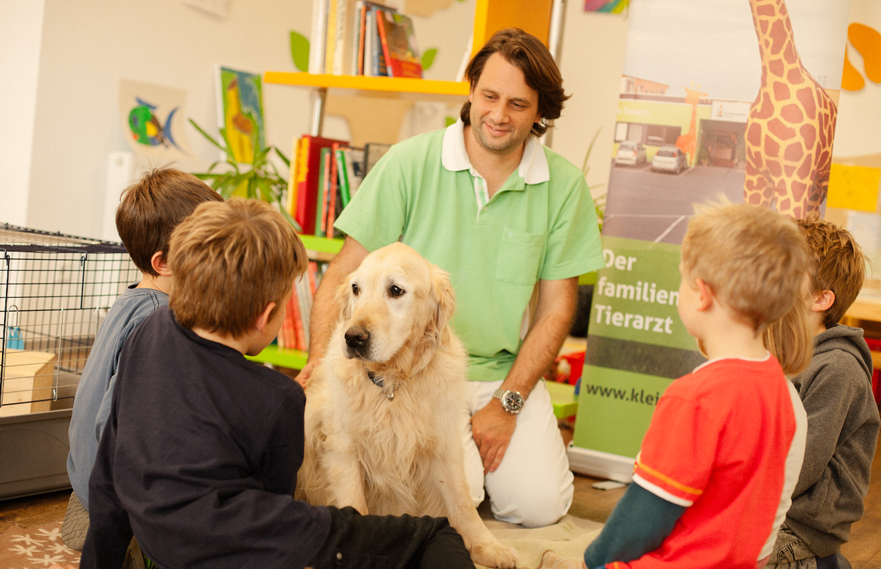 Ein Mann in einem grünen Poloshirt kniet mit einem Golden Retriever in einem Klassenzimmer mit Kindern, einem Bücherregal und einem Poster.