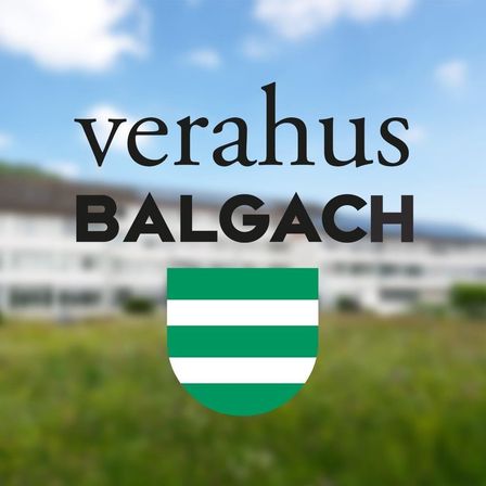Das Verahu Balgach-Logo zeigt ein Wappendesign mit grünen und weißen Streifen vor einem verschwommenen Hintergrund aus einem Grasfeld und einem bewölkten Himmel.