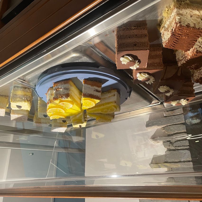 Eine Glasvitrine mit einer Drehplatte, die verschiedene Desserts wie Kuchen und Törtchen präsentiert, mit Reflexionen einer Steinmauer und Licht.