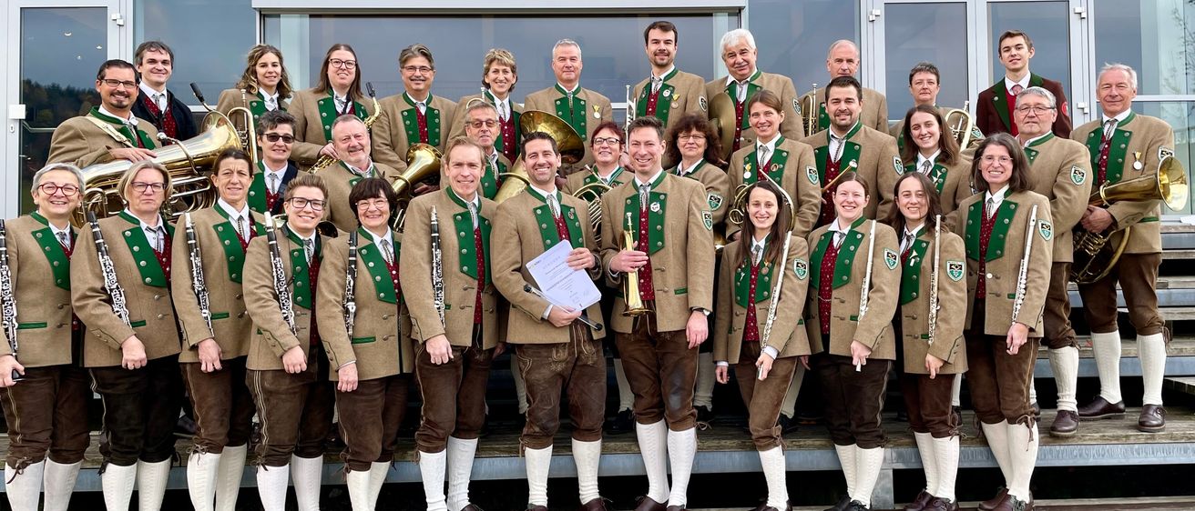 Eine Gruppe von Musikern in traditioneller deutscher Kleidung posiert für ein Foto. Sie halten verschiedene Instrumente wie Trompeten, Posaunen und Klarinetten. Sie stehen vor einem Gebäude mit großen Glasfenstern.