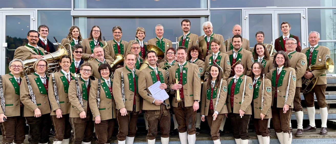 Eine Gruppe von Musikern in traditioneller deutscher Kleidung posiert für ein Foto. Sie halten verschiedene Instrumente wie Trompeten, Posaunen und Klarinetten. Sie stehen vor einem Gebäude mit großen Glasfenstern.