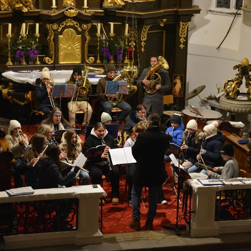 Ein Musikensemble spielt in einer Kirche mit rotem Teppich, verschiedenen Musikinstrumenten und einem Hintergrund aus goldenen Kerzenständern und Blumenarrangements. Der Dirigent steht vorne, und die Musiker sitzen und stehen.
