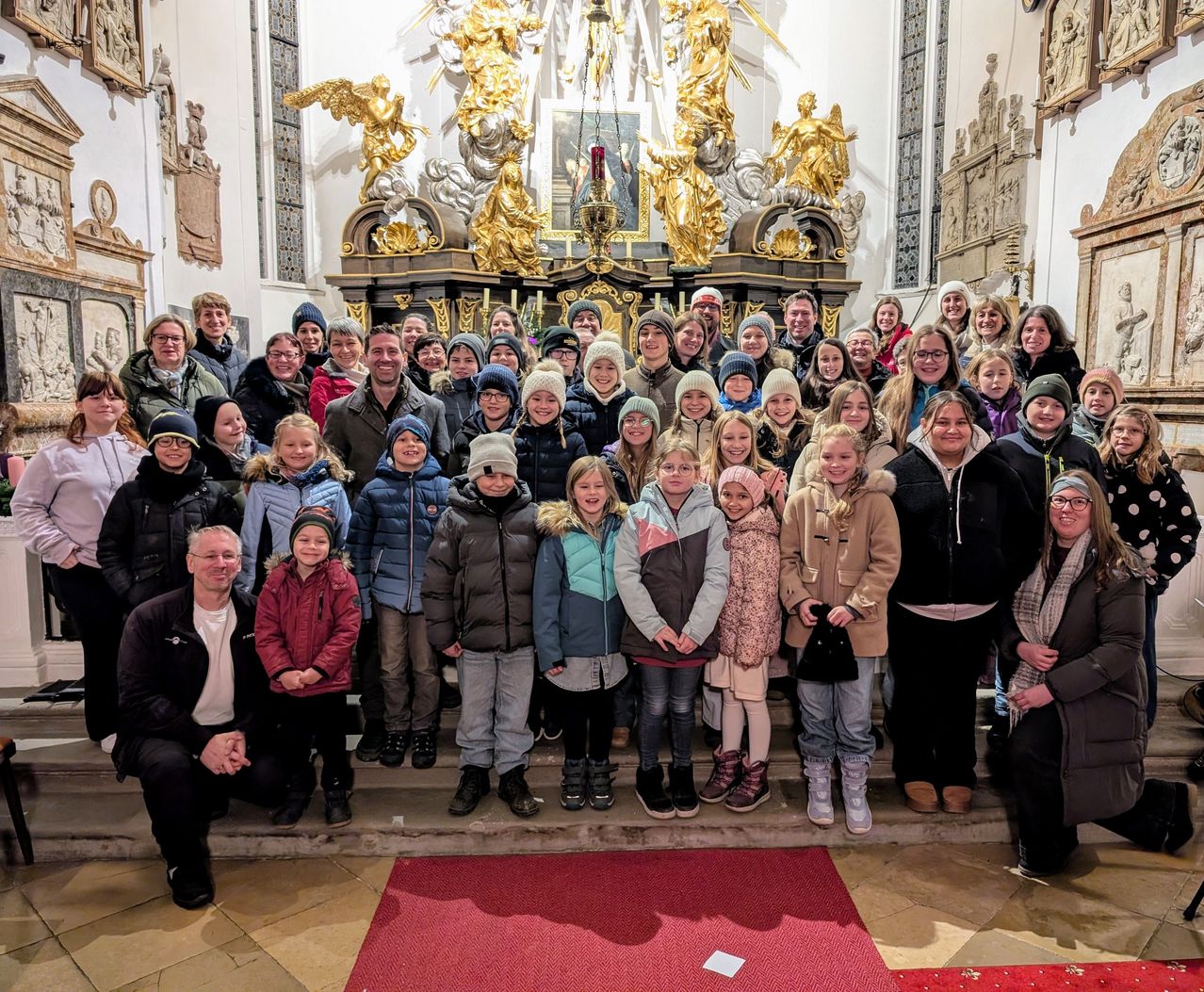 Eine Gruppe von Menschen, einschließlich Erwachsener und Kindern, posiert für ein Foto in einer Kirche mit kunstvollen Dekorationen. Sie stehen auf einem roten Teppich.