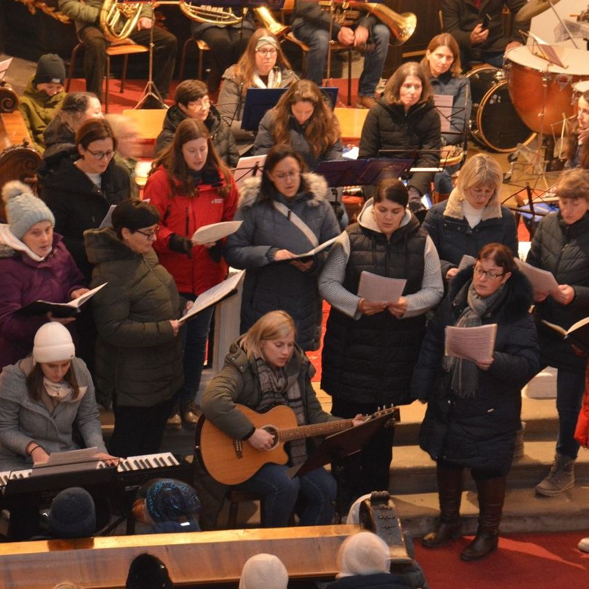 Ein Chor von Frauen in Wintermänteln steht in einer Kirche und singt. Dahinter spielen Musiker Instrumente wie Trommeln, Trompeten und Klavier.