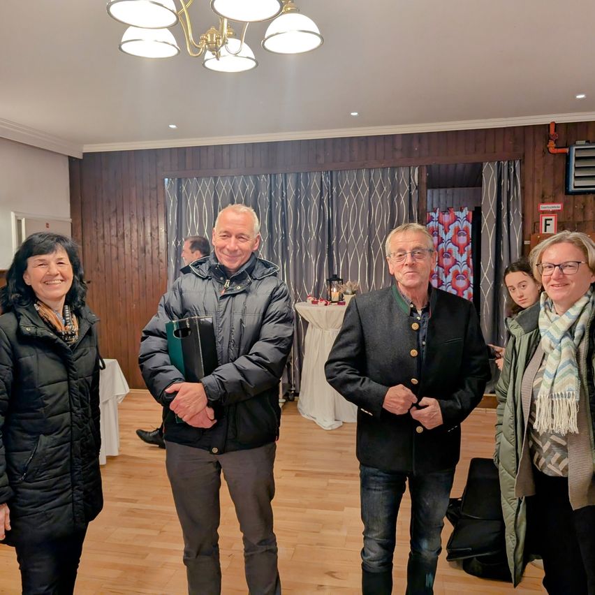 Four people stand indoors, smiling. Two men and two women. The room has wooden walls and a chandelier. Behind them, a table with a white cloth and a person in the background.