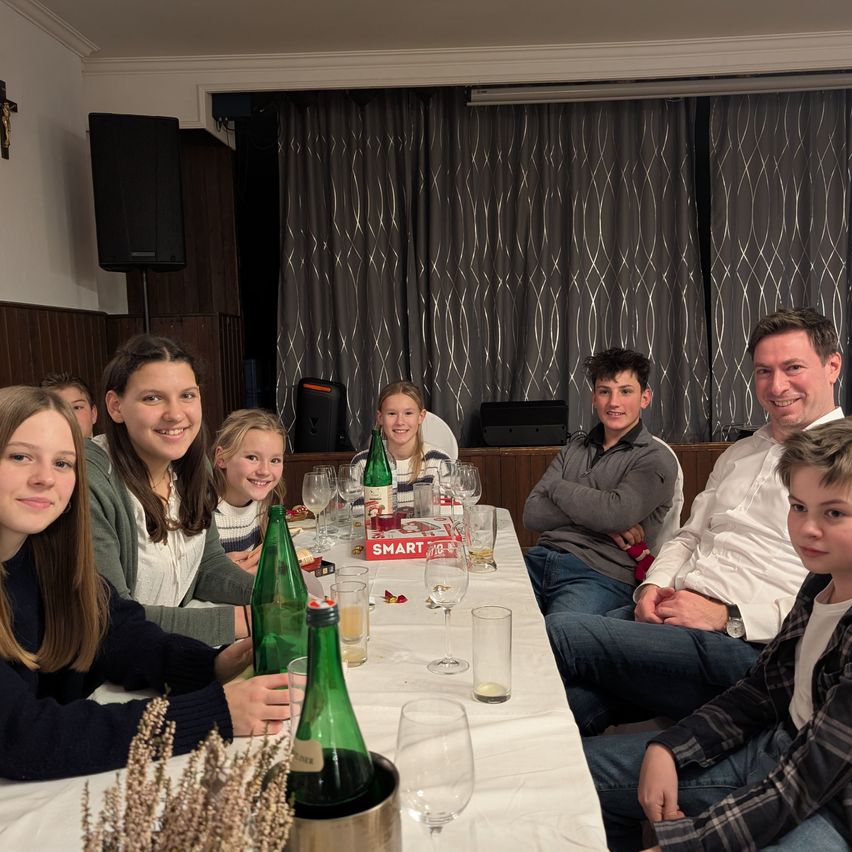 A family gathers around a table, smiling for a photo. Bottles and glasses are on the table, and a box labeled 'SMART' is visible. Two speakers and curtains are in the background.