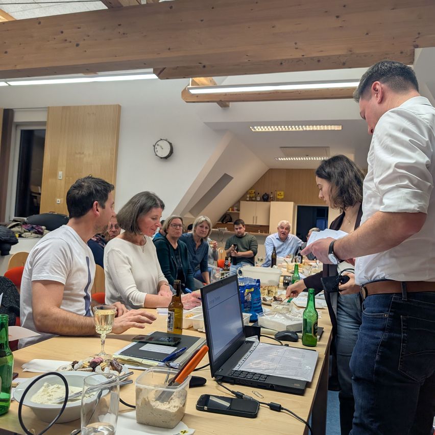 Eine Gruppe von Menschen sitzt um einen Tisch in einem Raum mit Holzbalken und diskutiert, genießt Essen und Getränke. Ein Laptop und Weingläser stehen auf dem Tisch.