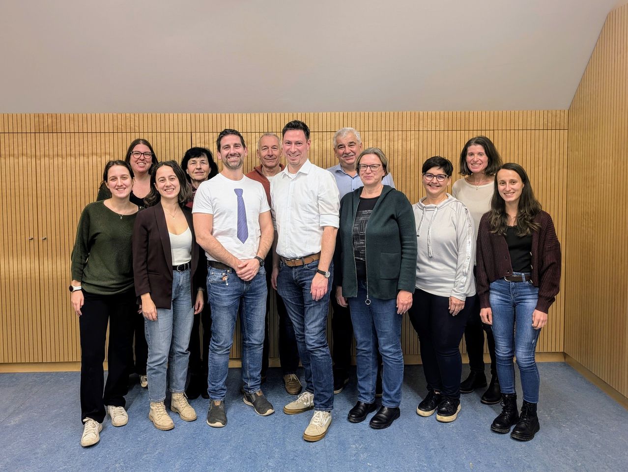 Eine Gruppe von Menschen, einschließlich Männer und Frauen, steht gemeinsam vor einer Holzwand. Sie alle lächeln und scheinen für ein Foto zu posieren.