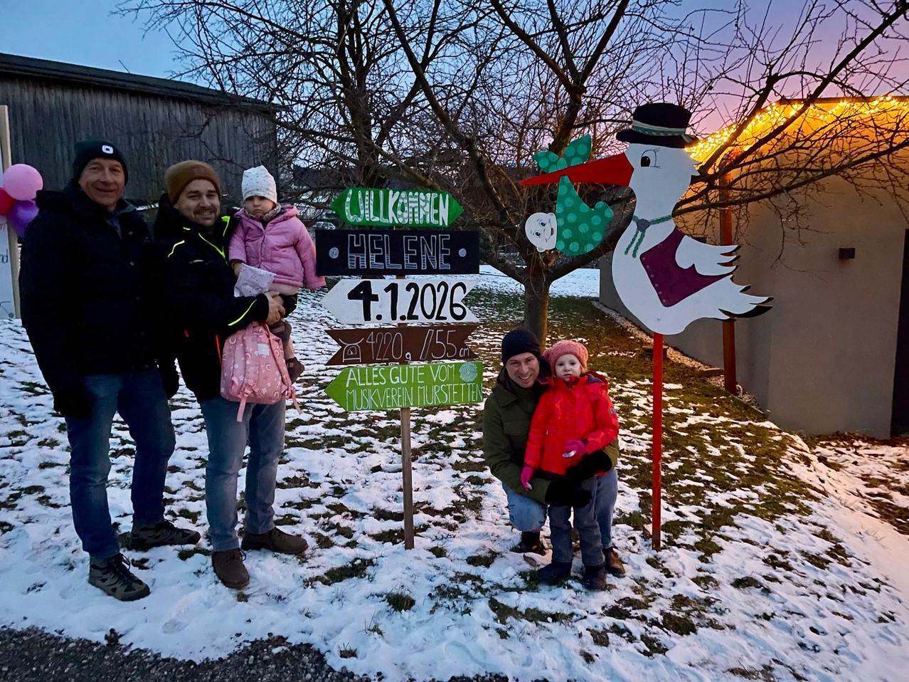 Vier Personen und zwei Kinder stehen im Schnee mit Schildern und einem Storch. Die Schilder lesen 'Helene' und '1.1.2026'. Das Kind auf der linken Seite hält eine Tasche. Das Kind auf der rechten Seite trägt einen Hut.