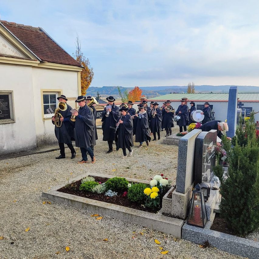 Eine Brassband spielt auf einem Friedhof. Sie tragen lange Mäntel und Hüte. Dahinter befindet sich ein Gebäude mit einem Fenster und einem Schild.