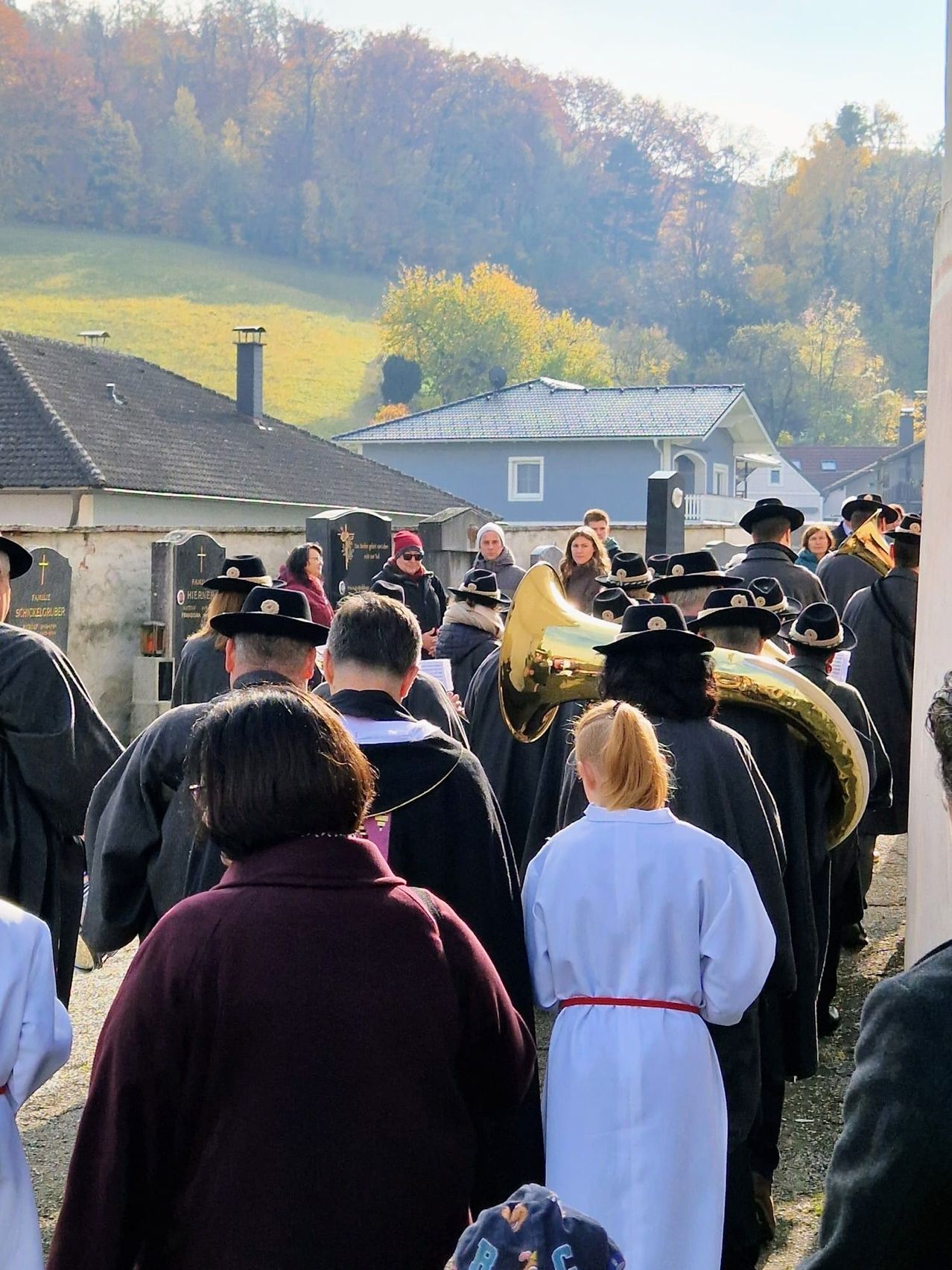 Eine Gruppe von Menschen, in Schwarz gekleidet, marschiert in einer Prozession. Einige halten Instrumente, und der Hintergrund zeigt eine ländliche Landschaft.