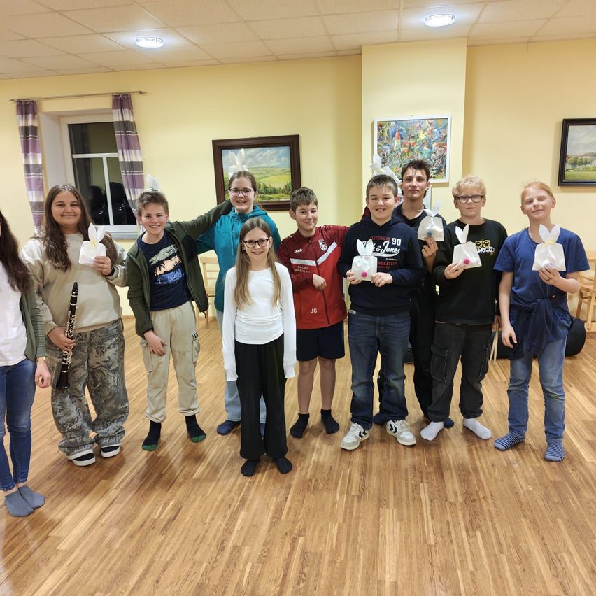 Eine Gruppe von Kindern und Jugendlichen posiert für ein Foto in einem Raum. Sie halten häschenförmige Tüten, und ein Kind hält eine Klarinette. Der Raum hat Holzböden und gelbe Wände.