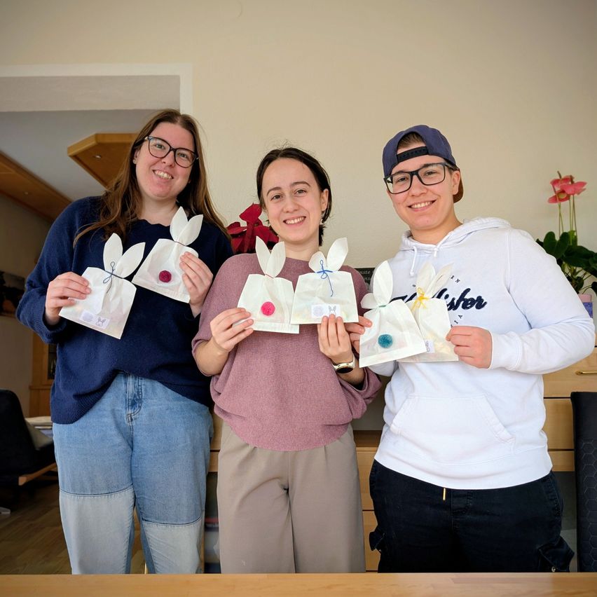 Drei Menschen posieren mit Osterdekorationen in einem Raum. Einer hält einen Papierbeutel mit einem Hasen, ein anderer hat einen ähnlichen Beutel und der Dritte hält zwei Beutel.