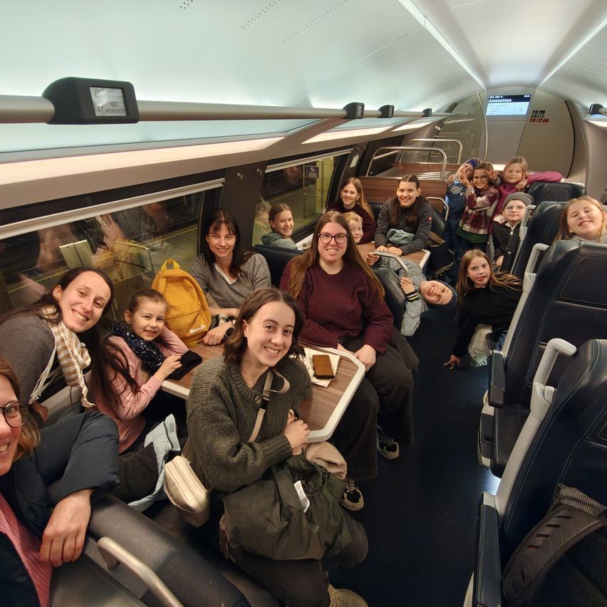 Eine Gruppe von Menschen sitzt in einem Zug. Einige lächeln, während andere in die Kamera schauen. Eine Frau in der Mitte hält eine Serviertablett mit Essen.