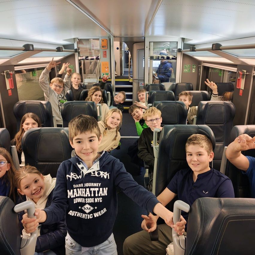 Eine Gruppe von Kindern in einem Zug, die lächeln und für ein Foto posieren. Einige tragen Brillen, ein Junge trägt ein Manhattan-Sweatshirt und ein anderer hält einen Griff.