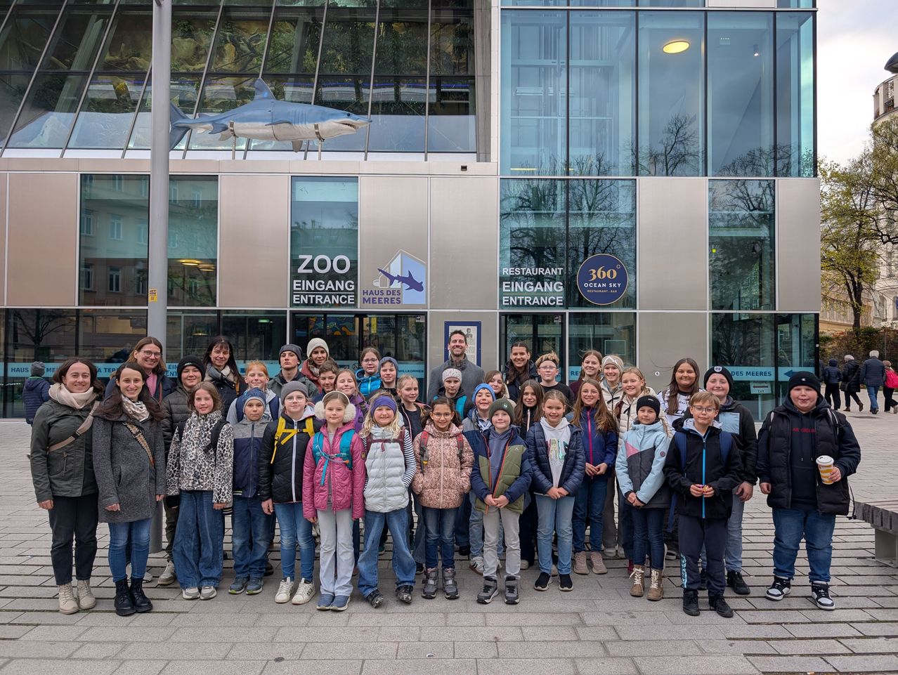 Eine Gruppe von Schülern posiert für ein Foto vor einem Gebäude mit einer Haifischskulptur darüber. Das Schild lautet 'ZOO EINGANGE ENTRANCE' und 'RESTAURANT EINGANGE ENTRANCE'.