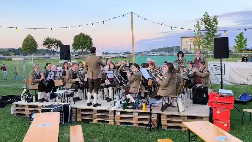 An orchestra is performing on an outdoor stage. They are wearing uniforms. They are playing different instruments. There are speakers on both sides of the stage.