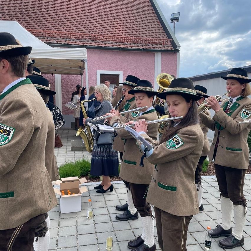 A group of people in traditional attire are playing instruments in front of a pink building. They are holding flutes and other brass instruments.