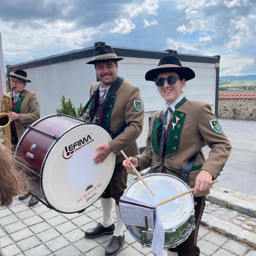 Three musicians, two playing drums and one playing saxophone, dressed in traditional German attire, are performing outside.