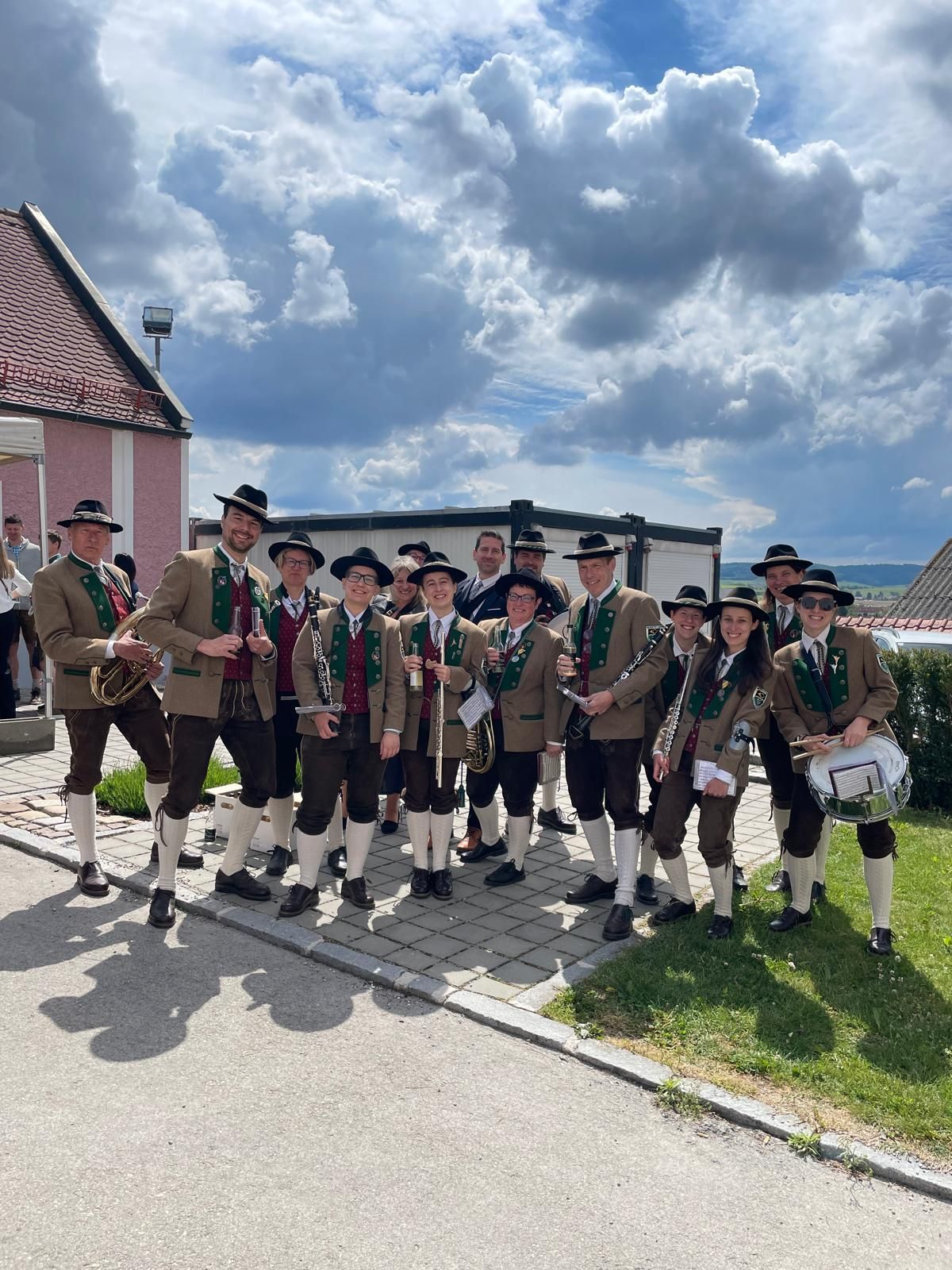 A group of musicians dressed in traditional Bavarian attire poses for a photo. They are holding various musical instruments. The photo was taken on a sunny day.