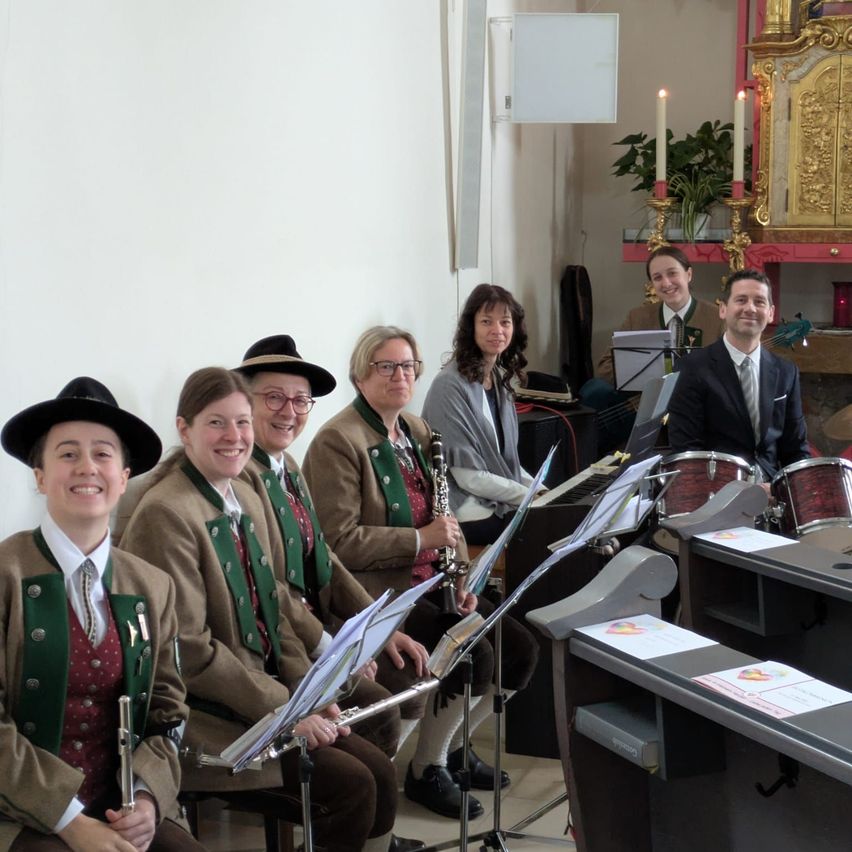 A group of musicians in traditional attire, seated with their instruments, poses for a photograph in a room with a piano and candles.