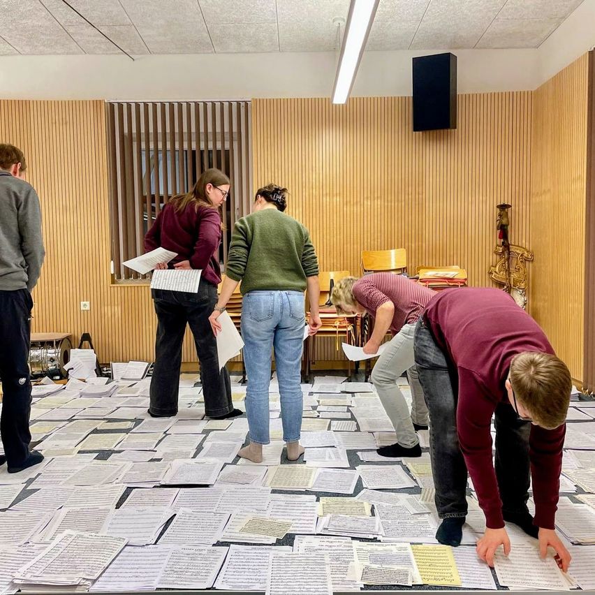 Fünf Personen in einem Raum, stehend auf verstreuten Musikpartituren, halten Papiere in den Händen. Ein Lautsprecher ist an der Wand montiert, und ein Holzobjekt befindet sich in der Nähe.
