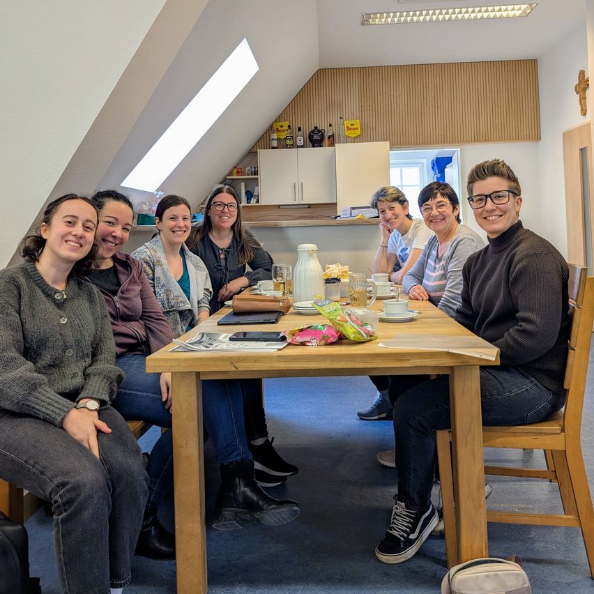 Eine Gruppe von Frauen sitzt um einen Tisch in einem Raum. Eine Frau lächelt. Der Tisch hat eine Karaffe, Tassen und einen Teller. Dahinter ist eine Küche mit Schränken und einem Fenster.