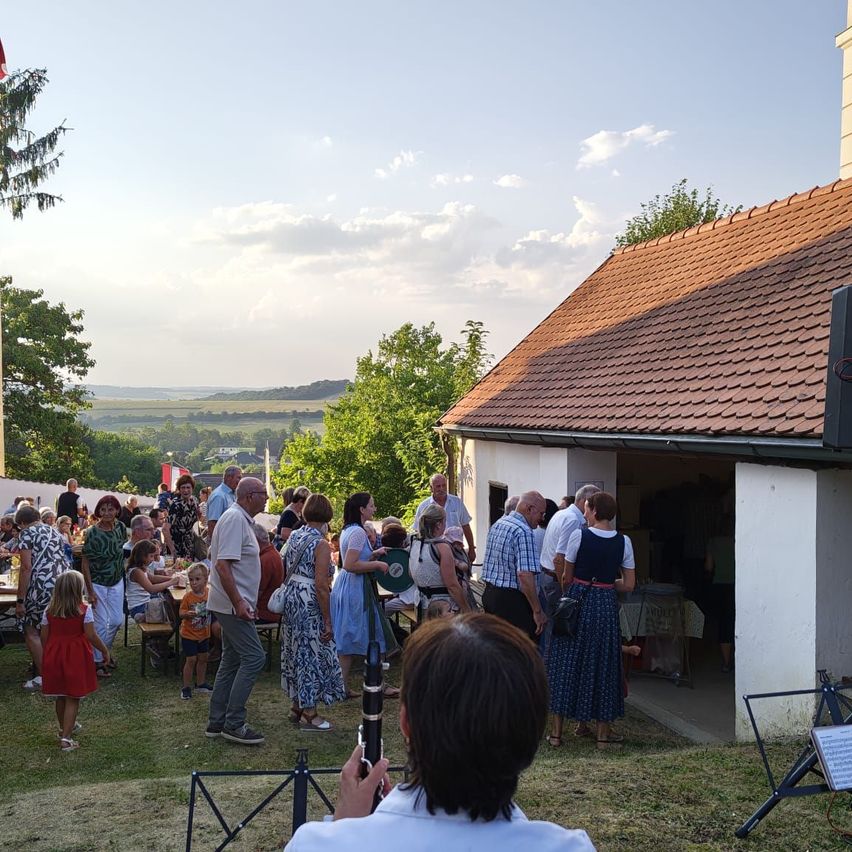 Eine Gruppe von Menschen versammelt sich auf einem Rasen, möglicherweise für eine Gemeinschaftsveranstaltung. Einige stehen, andere sitzen an Tischen. Ein Gebäude mit einem roten Dach ist im Hintergrund zu sehen.