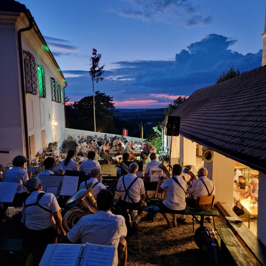 Eine Blaskapelle spielt auf einer Bühne vor einem Gebäude mit grünen Lichtern, unter einem Dämmerungshimmel. Das Publikum sitzt auf Bänken und Stühlen und genießt die Musik.