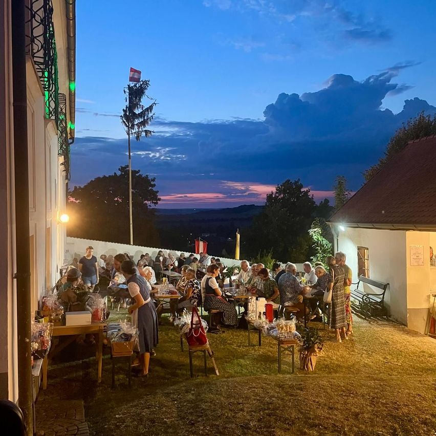 Eine Gruppe von Menschen versammelt sich im Freien bei Dämmerung, sitzt um Tische mit verschiedenen Gegenständen herum, unter einem klaren blauen Himmel mit einigen Wolken.