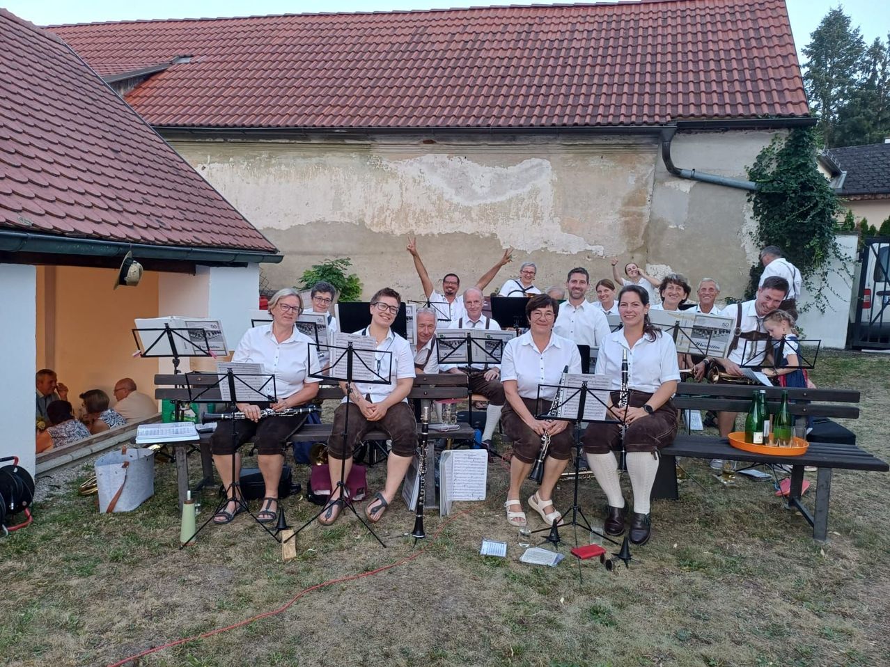 Eine Gruppe von Musikern in traditioneller Kleidung sitzt auf Bänken im Freien, mit Musikinstrumenten und Notenblättern vor ihnen. Die Umgebung scheint ein ungezwungenes Open-Air-Konzert zu sein.