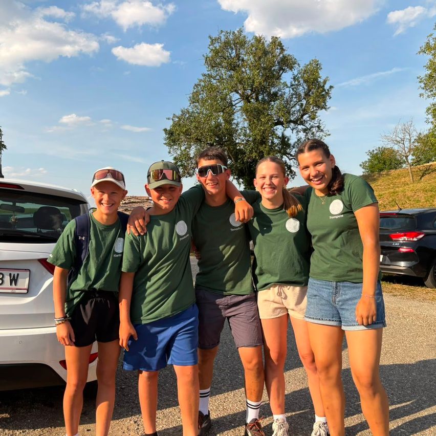 Five young people in matching green shirts pose for a photo in a parking lot with a white car and trees in the background.