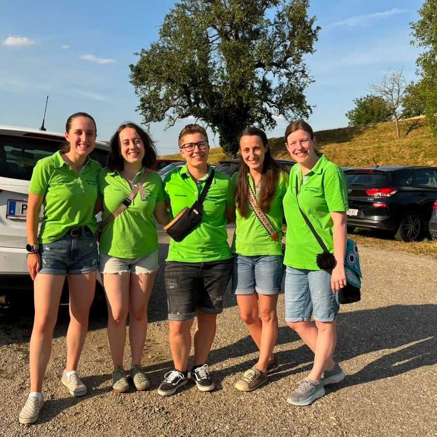 Five women in green shirts and denim shorts pose for a photo in a parking lot with a tree and parked cars in the background.