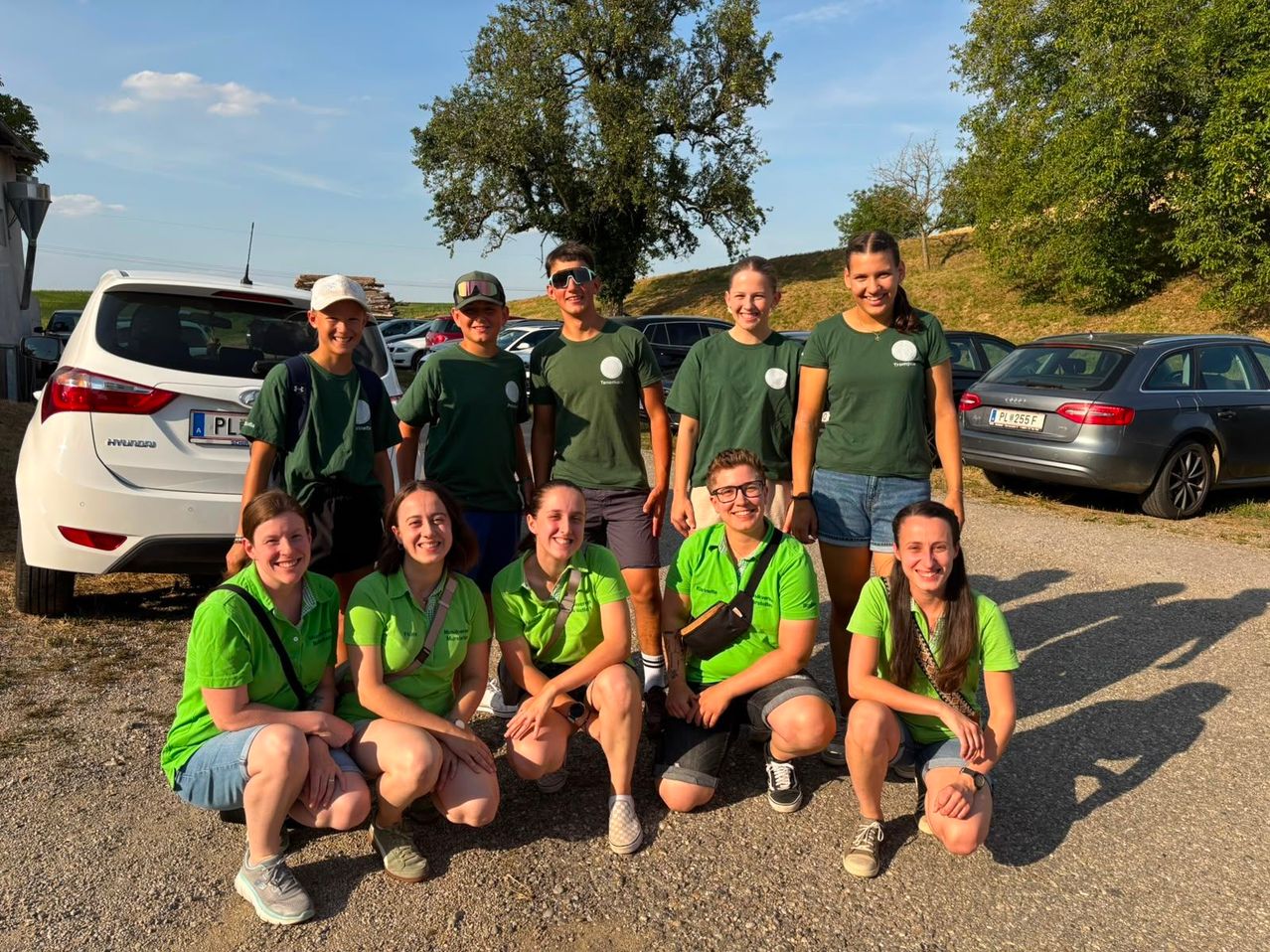 A group of young people in green shirts are posing for a photo in a parking lot with cars and trees in the background.
