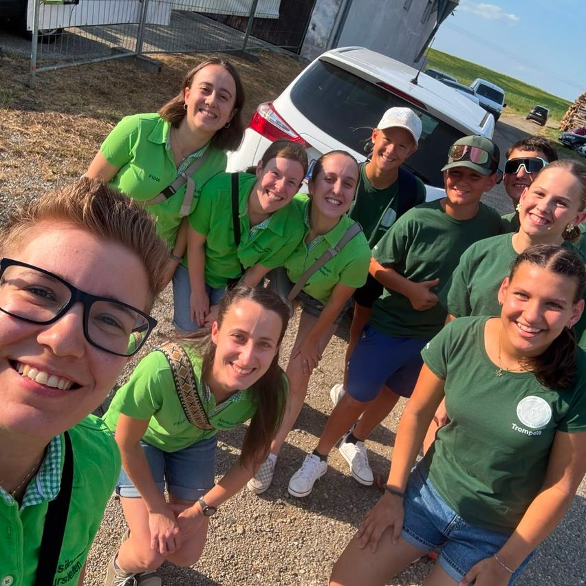 A group of young people in green shirts are smiling and posing for a photo outside. They are standing in front of a white car and a fence.