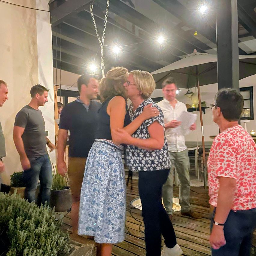 Eine Gruppe von Menschen versammelt sich auf einer Holzterrasse, zwei Frauen umarmen sich, während andere zusehen. Ein Mann hält ein Stück Papier.