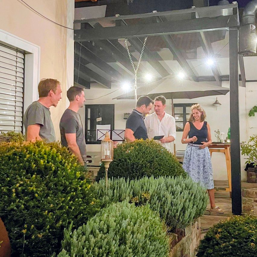 Fünf Personen stehen nachts in einem Garten, einer Mann hält ein Tablet. Eine Frau in einem Blumenrock steht bei einem Tisch mit einer Lampe.