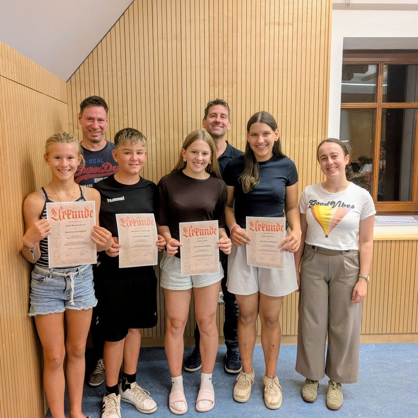 A group of young individuals, including both boys and girls, are standing together and holding certificates. They are smiling and posing for a photo. Behind them, two men are standing and smiling as well. They are in a room with wooden walls and a blue floor.