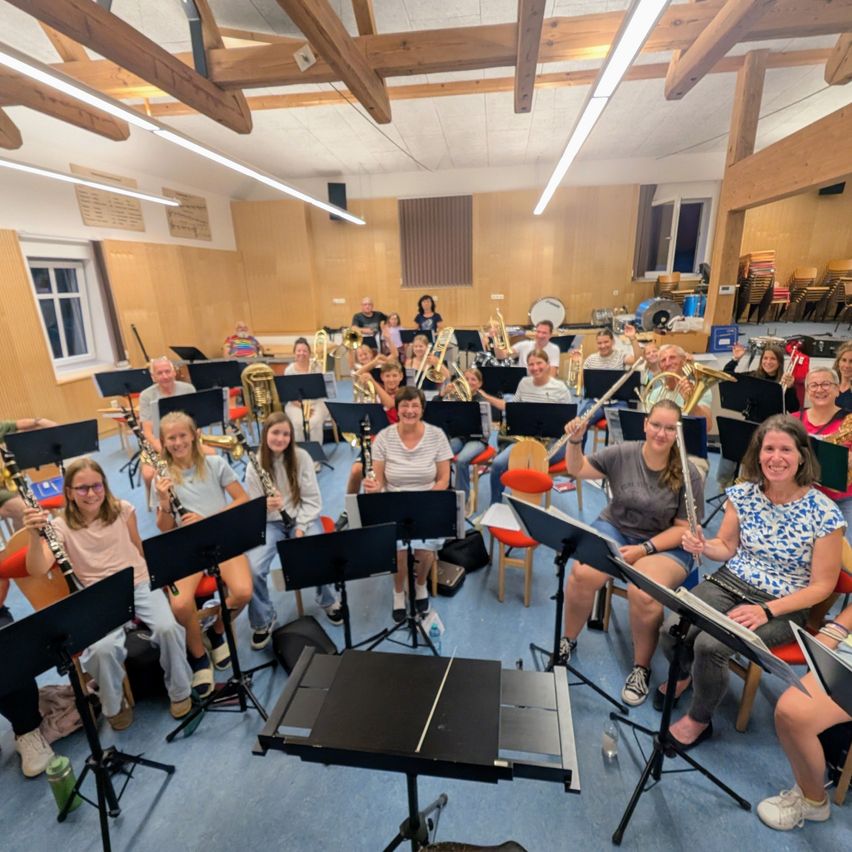 A group of musicians, both adults and children, are sitting in a room with musical instruments, possibly preparing for a rehearsal or performance.