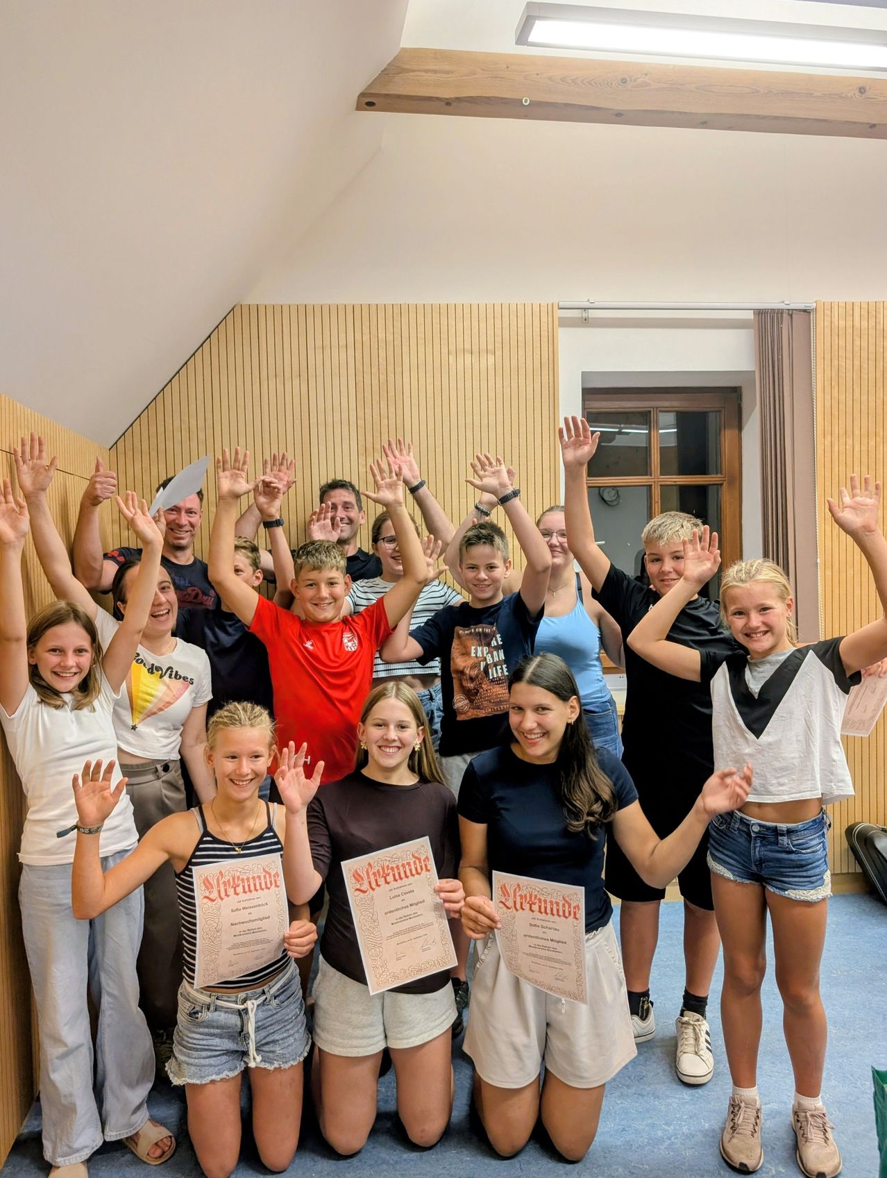 A group of young people and adults are posing for a photo with certificates in their hands. They are smiling and raising their hands. The room has wooden walls and a window.