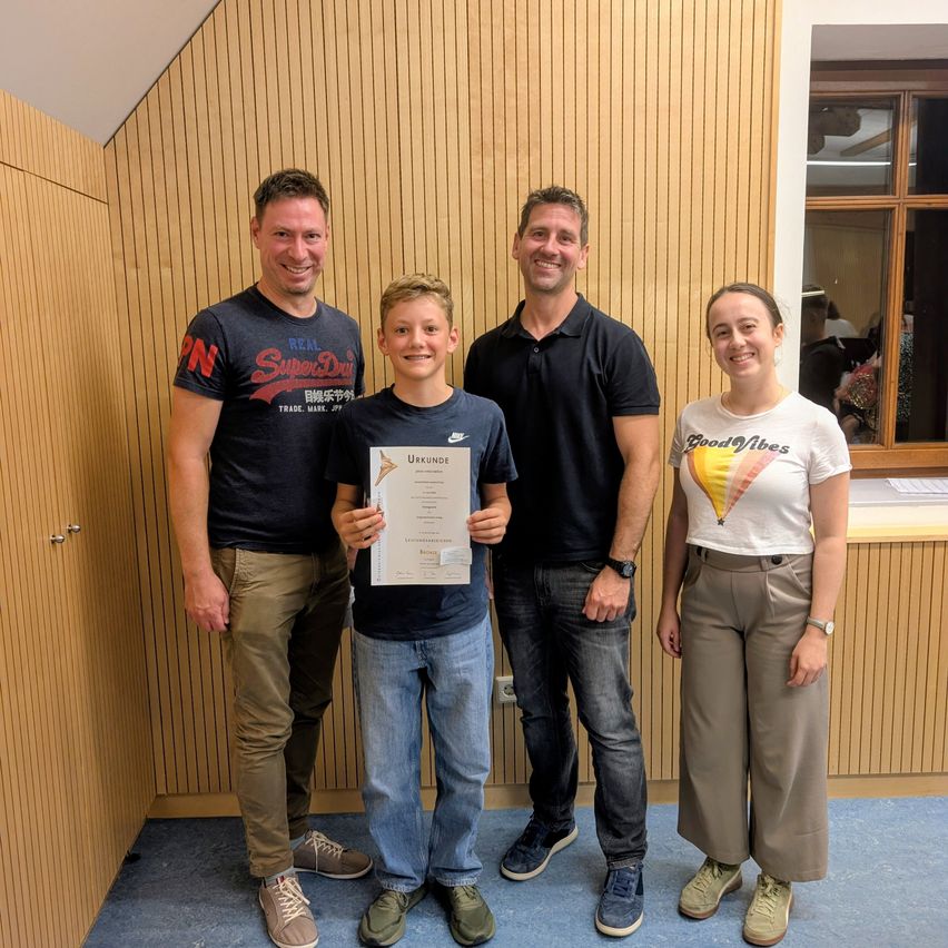 Four people stand together in a room, smiling for a photo. A boy in the center holds a certificate. The man on the left wears a shirt with a logo, and the man on the right wears a black polo. A woman stands to the right, wearing a white shirt with a balloon design.