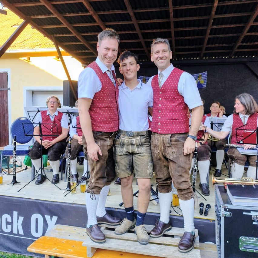Three men in traditional Bavarian attire stand on a stage, smiling for a photo. Behind them, musicians play various instruments, and a banner with text is visible on the stage.