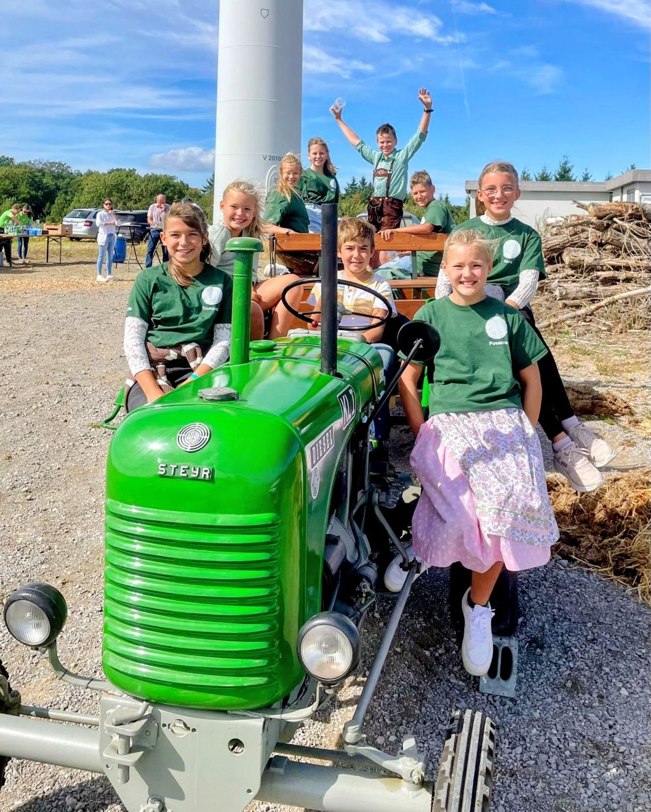 A group of children in green shirts sit on a green tractor, with some standing behind it. They are smiling and posing for a photo. In the background, there are trees, a white building, and a pile of wood.