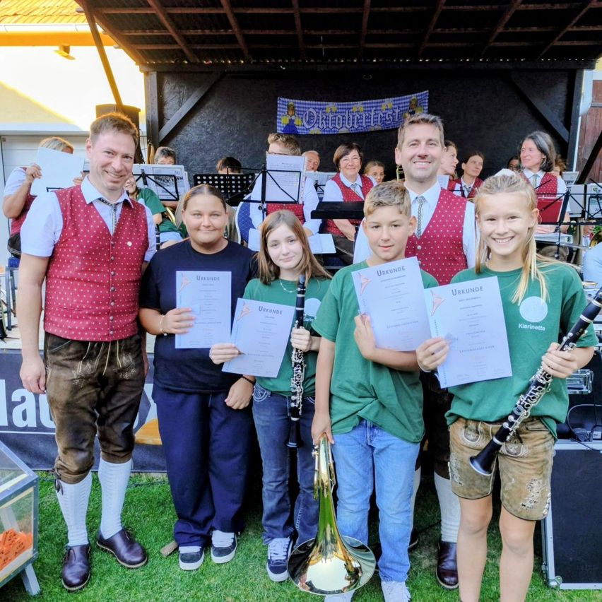 A group of children and two men stand on a stage holding certificates and musical instruments. Behind them, a band is playing on stage.