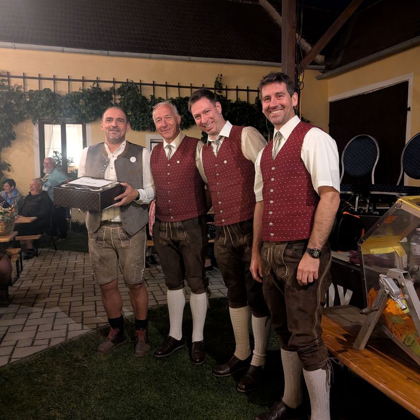 Four men dressed in traditional German attire, standing together and smiling for a photo. One man holds a box. Behind them, a building with a patio and chairs.