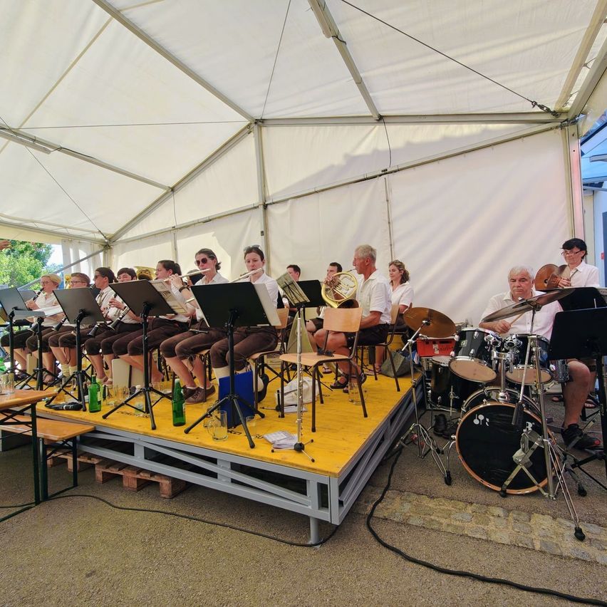 Eine Brassband spielt unter einem weißen Zelt, wobei mehrere Musiker, darunter ein Mann, der eine Trompete spielt, auf einer Bühne sitzen.