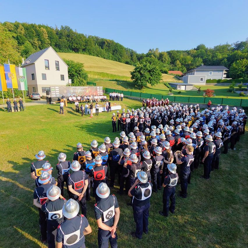 Eine große Gruppe von Menschen in Uniformen und Helmen versammelt sich auf einem Feld für eine Veranstaltung, mit Häusern und Bäumen im Hintergrund.