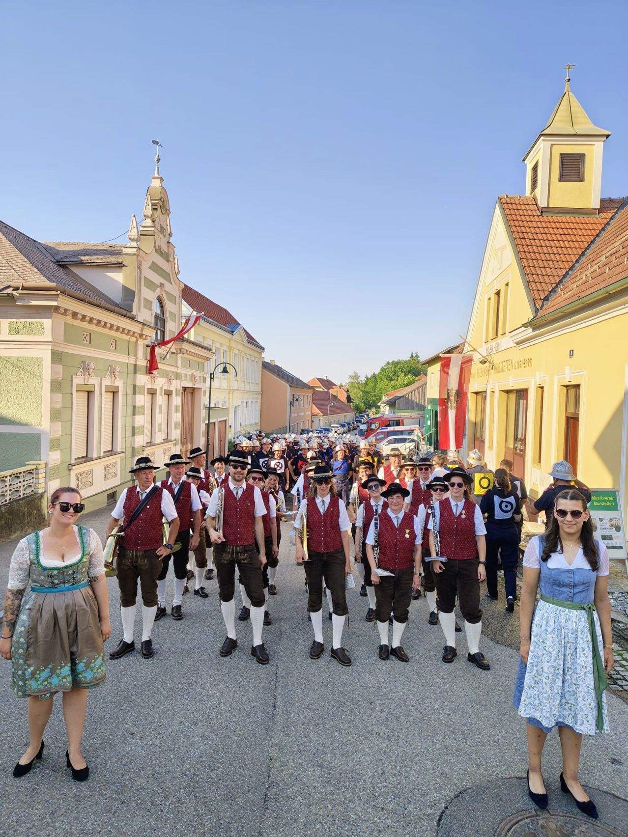 Eine Band in traditioneller Kleidung marschiert die Straße entlang, Zuschauer schauen zu. Gebäude säumen die Straße, und eine Flagge weht auf einem Pfahl.